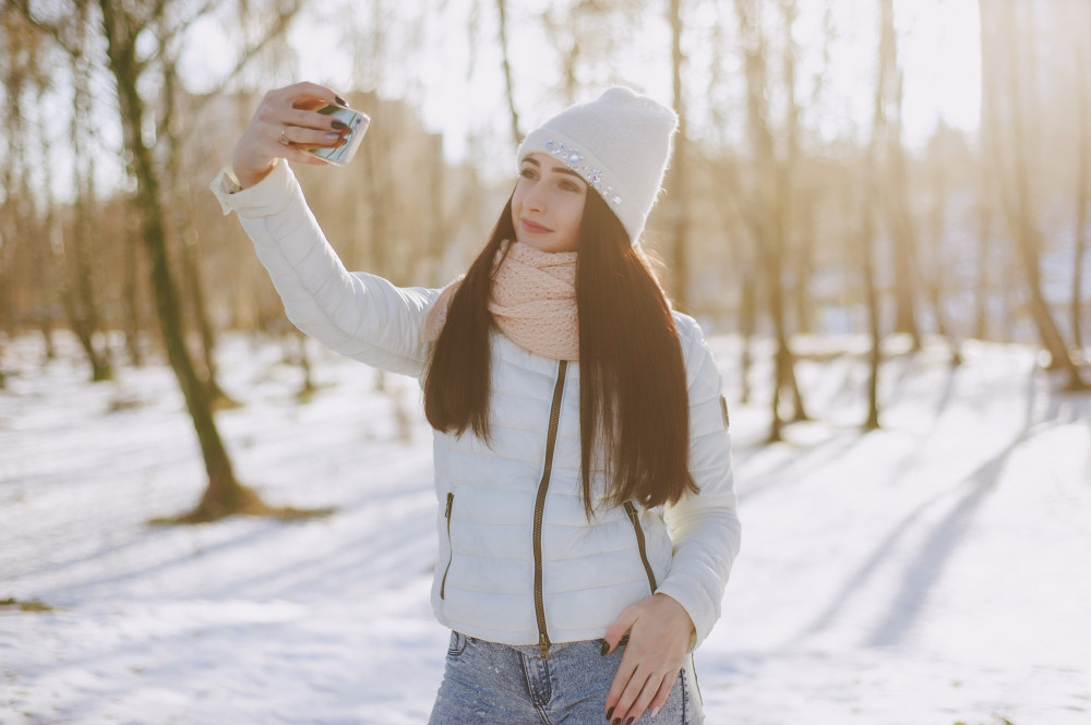 Девушка в белой куртке делает селфи на фоне зимнего пейзажа.
